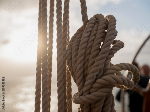 Foto Twisted natural fibre ropes hang in warm backlight on the deck of a sailing ship, fibres glowing against the shining water