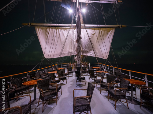 An empty passenger deck at night with wicker chairs and tables under a square sail lit from below, harbour lights far off.