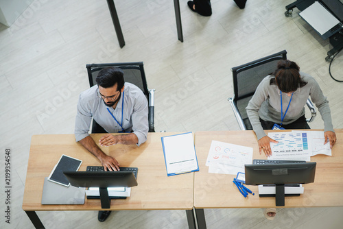Top view of business professionals working at computer workstations and reviewing charts, reports, and digital information in a modern office.