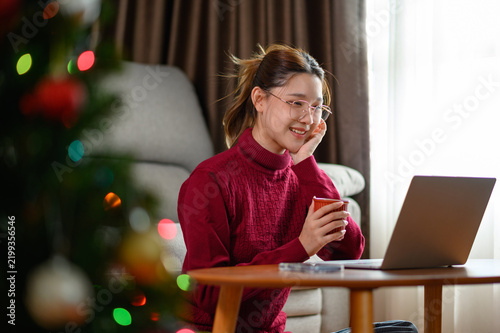 Smiling young Asian woman working remotely from home with a laptop while enjoying a cup of coffee during Christmas holiday. Cozy festive workspace, remote work,  holiday lifestyle concept.