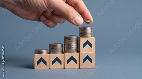 Hand adding coin to growing stack of money blocks with upward arrows
