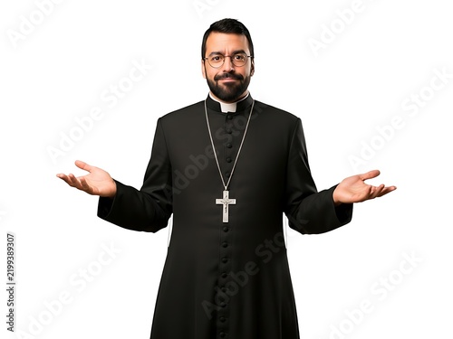 A bearded priest in a black cassock with a cross necklace gesturing openly