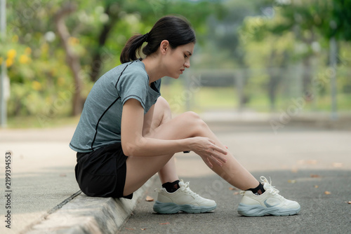 Obraz na plátně Young woman sitting on curb with leg pain after running in park, holding sore shin muscle, sports injury and overuse strain outdoor fitness concept