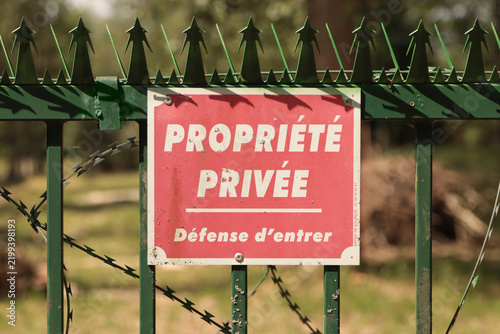 Fotografie A rectangular red sign with white text is attached to a green metal spiked fence