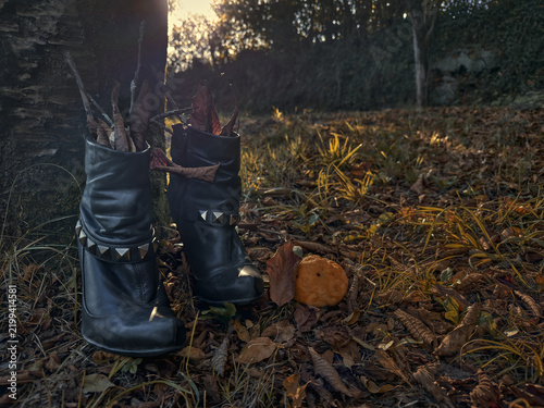 Black leather witch boots filled with dry leaves and branches beside a small orange pumpkin in an autumn woodland, with atmospheric backlight and generous copy space for a Halloween concept.