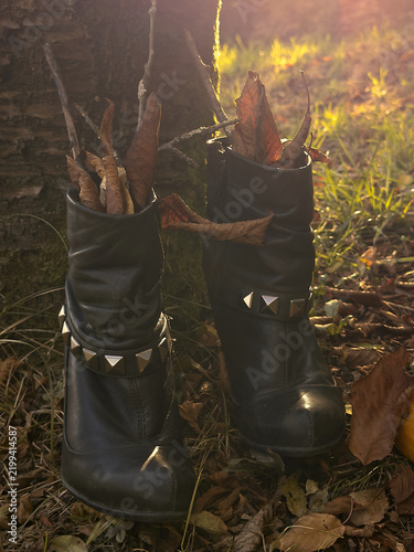 Black leather witch boots filled with dry autumn leaves and branches beside a tree, illuminated by warm golden backlight in a mysterious woodland Halloween scene.