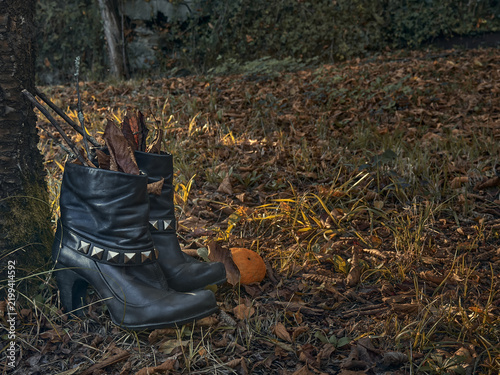 Black leather witch boots filled with dry leaves and branches beside a small pumpkin in an autumn woodland, arranged on the left with generous copy space for Halloween advertising and seasonal designs