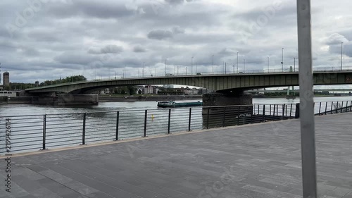 View of the Rhine River embankment in Cologne with a bridge, cargo barge ship, and overcast cloudy sky background.