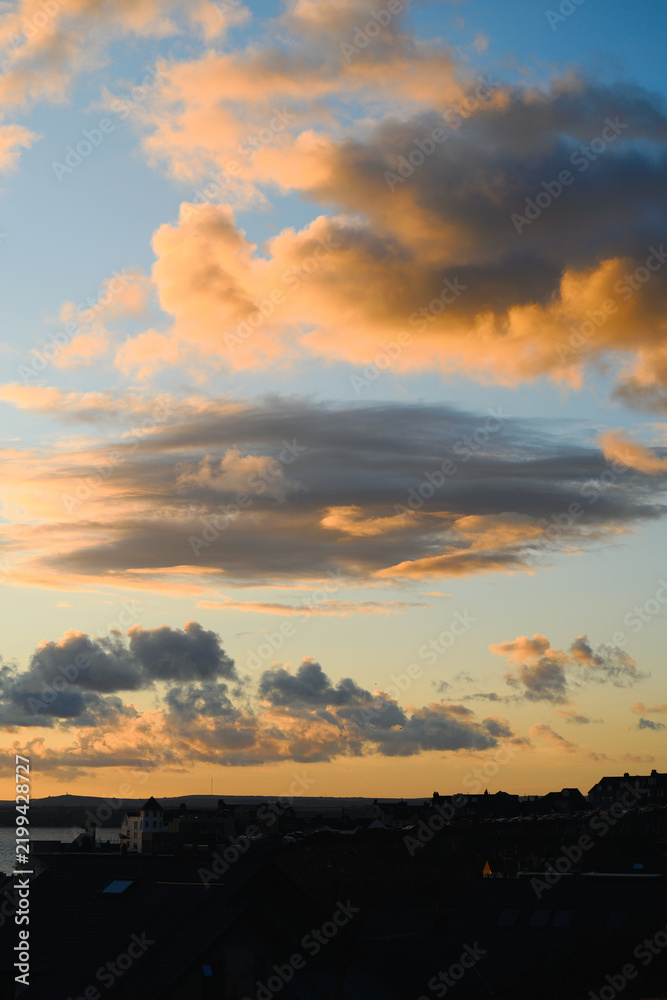 Obraz premium Glowing sunset clouds drift above coastal rooftops in Saint Ives, Cornwall, England, with warm evening light, layered sky and distant shoreline view.
