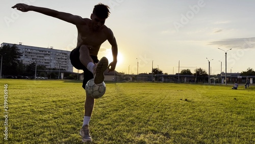 Young man juggling soccer ball on stadium at sunset. Professional footballer kicking ball at green field. Sportsman practicing tricks at meadow with sunlight at background. Freestyle football