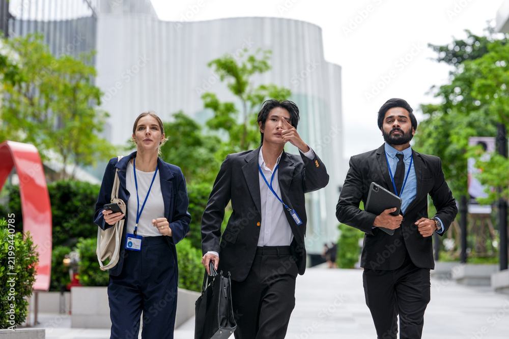 Fototapeta premium Three young business professionals wearing conference badges advance together down the corporate promenade along a sunlit walkway between contemporary corporate (frame 146)