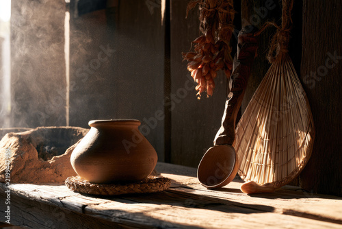 Traditional rural kitchen hearth with earthenware pot, woven trivet, dried garlic, shallot, spoon, fan, smoke and warm sunlight