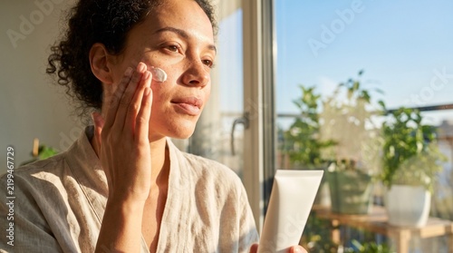 Jovem e bela mulher brasileira aplicando creme facial hidratante perto da janela, rotina de cuidados com a pele.