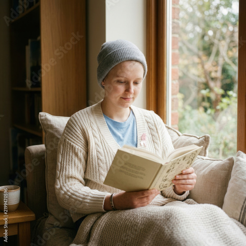 Young Woman Wearing Beanie Reads Book Comfortably By Window