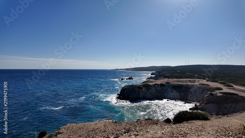 Foto Harsh cliffs rise above the deep blue sea, where waves crash against rocks and the coastline stretches into the distance under a clear sunny sky