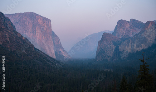 View of El Capitan and the granite cliffs of Yosemite Valley at dusk with hazy sky and dense forest in Yosemite Valley, California, United States.