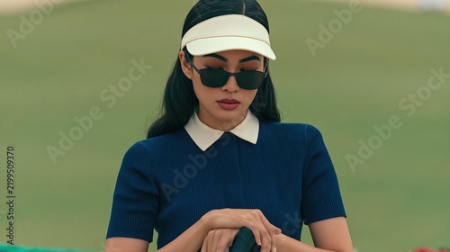 A stylish woman wearing sunglasses and a visor, dressed in a navy shirt with a white collar, is concentrating while holding a golf club on a green course.