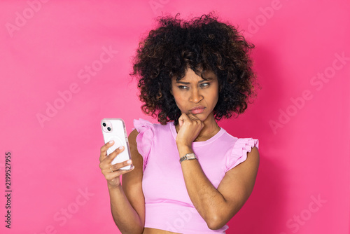 young latina woman with an afro hairstyle looking at her mobile phone with suspicion or doubt