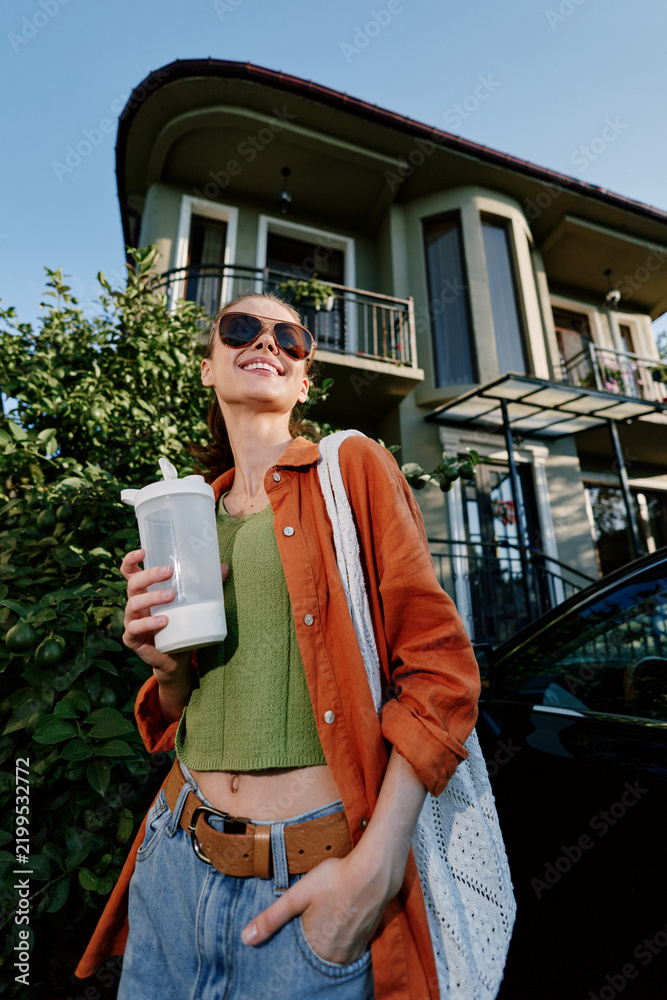 Obraz premium Young woman in an orange jacket smiles while holding a cup outdoors in front of a modern house, wearing sunglasses, a green top, jeans, and a tote bag.