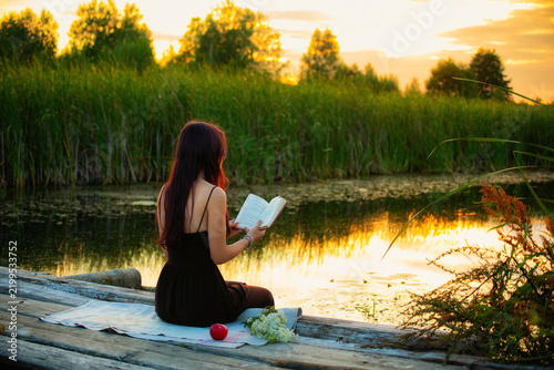 Young woman reading a book on a wooden pier by a calm lake during a beautiful golden sunset