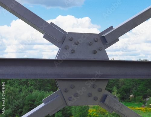 Cuadro en lienzo Detailed view of a bolted steel framework connection featuring diagonal beams and a central gusset plate, set against a background of green forest and cloudy blue sky