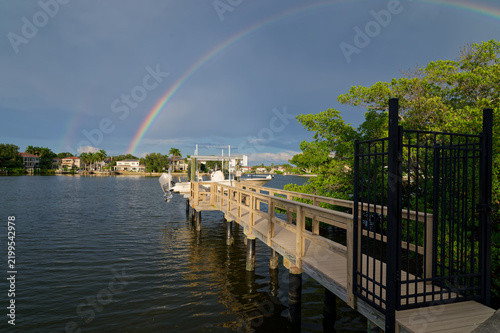 A vivid rainbow arches over a residential waterfront marina with a wooden dock, moored boats, lush mangroves and reflective water Coffee Pot Park bay in St. Petersburg, FL conveying serenity, hope and