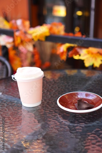 Takeaway coffee cup with chocolate candy on glass table of street cafe with autumn leaves decor. Self-care, cozy fall break and Halloween season concept. Warm seasonal atmosphere outdoors.