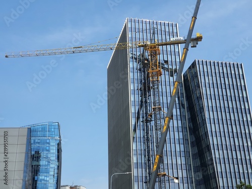 Modern urban construction site featuring high-rise glass office buildings under a clear blue sky. A prominent yellow tower crane is mounted on the facade of the tallest building, displaying a