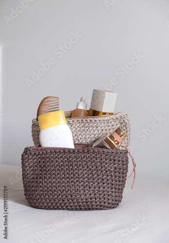 Two textured crocheted pouches in beige and mauve containing care products and a comb placed on a bed with beige bedding and a light-colored surface.