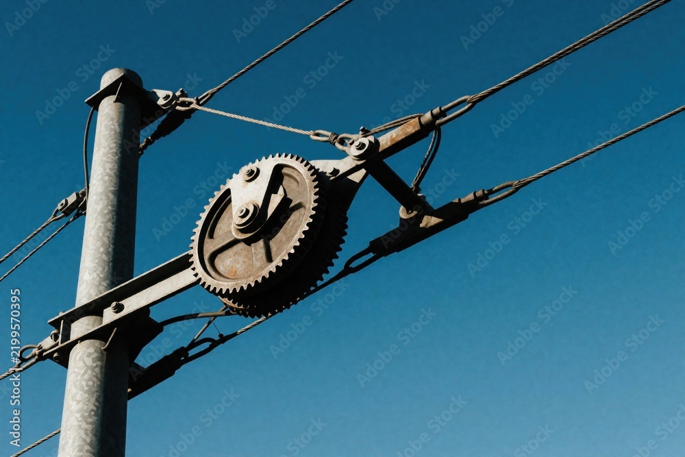 Obraz premium Overhead tramway cable pulley, metal support pole against clear blue sky. Industrial gear mechanism on electric line provides tension for urban transit. Technical engineering equipment connects wires