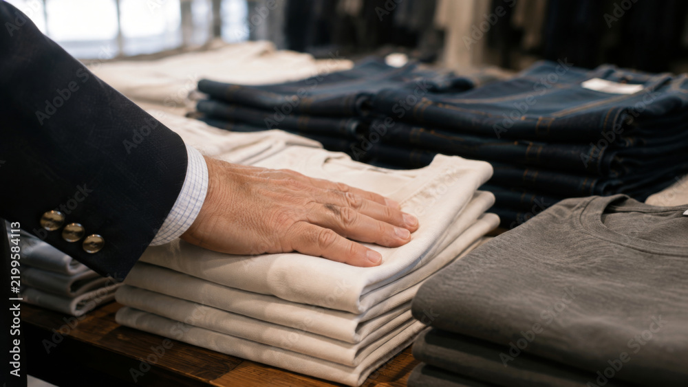 Fototapeta premium Customer touching a stack of neatly folded pants in a clothing store with jeans and shirts on display, representing retail shopping and casual fashion
