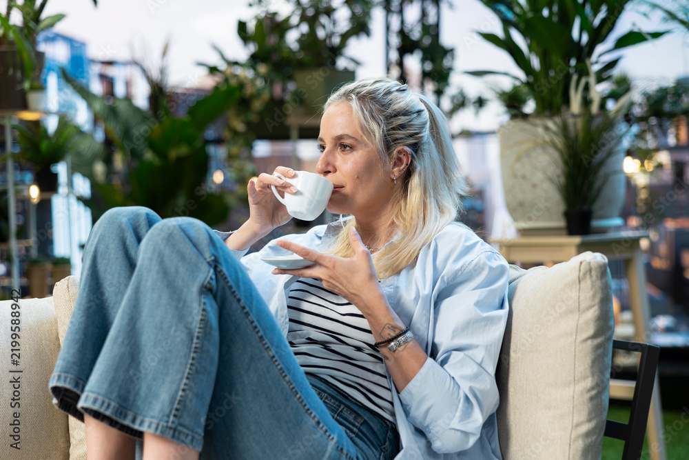 Fototapeta premium Woman drinks coffee from a white cup while relaxing on a rooftop garden sofa, enjoying a quiet leisure moment among plants in an urban setting.