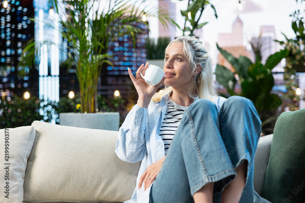 Fototapeta premium Woman drinks coffee from a white cup while relaxing on a rooftop garden sofa, enjoying a quiet leisure moment among plants in an urban setting.