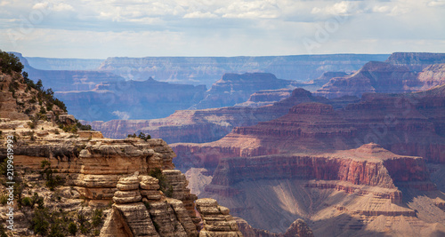Layered Grand Canyon cliffs and mesas displaying colorful sedimentary rock strata beneath clouds in a broad Arizona national park landscape