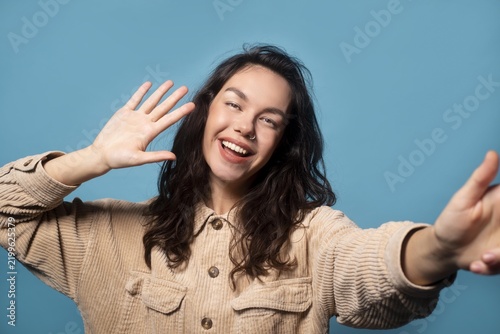 фотографія Smiling Young Female With Light Skin Gesturing While Waving Hand At Camera, Displaying Happiness In Studio Setting With Blue Background