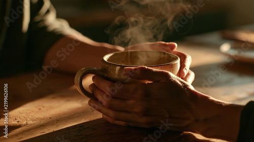 Warm Embrace: Two hands gently cradle a steaming mug of hot beverage, casting a warm glow on a rustic wooden table, evoking comfort and cozy intimacy.