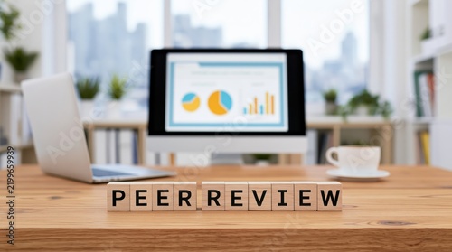 Peer review process with wooden blocks on a desk near a computer monitor and laptop for scholarly research analysis