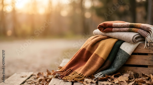 A stack of cozy wool blankets rests on a wooden bench amidst fallen autumn leaves in a sunlit park during sunset.