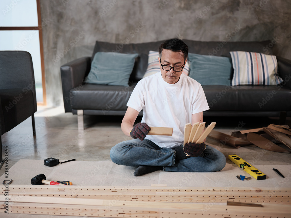 Fototapeta premium Asian mature man sitting and preparing flat-pack furniture parts and DIY equipment for self-assembly, home improvement, wooden craft installation, hobby workshop, carpentry handyman active lifestyle.