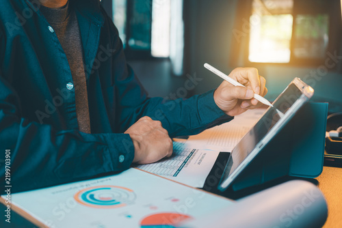 Businessman hand touching tablet screen with stylus pen, analyzing financial report document on desk, digital workspace and business concept.