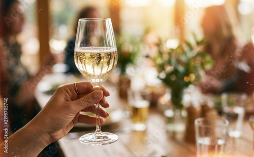 Close up of a female hand holding a glass of sparkling white wine at a festive dinner party with blurred background
