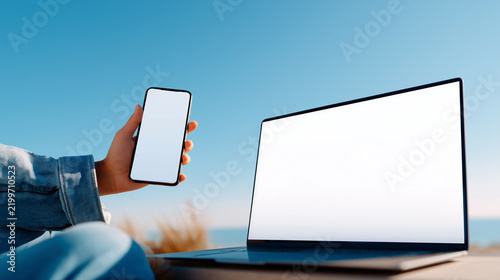 Hand holding blank smartphone next to laptop with white screen