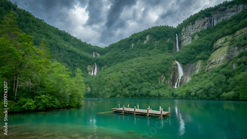 A wooden dock floating on a calm turquoise mountain lake, surrounded by steep cliffs covered in dense green forest and multiple cascading waterfalls under a dramatic, cloudy, and overcast sky