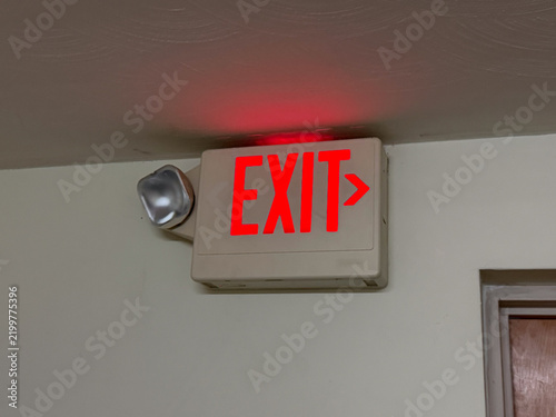 Cuadro en lienzo Red illuminated EXIT sign with arrow and emergency light fixture mounted on a light-colored wall, symbolizing safety, evacuation, and critical guidance in a building interior