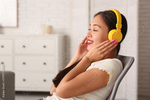Beautiful young happy Asian woman in headphones listening music on chair at home