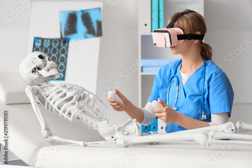 Female doctor in VR glasses with controllers and human skeleton model on couc...