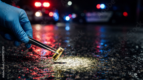 Crime scene investigator collecting bullet casing with tweezers on wet asphalt at night, forensic evidence investigation concept.