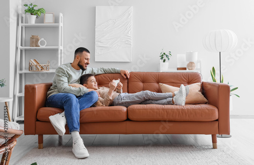 Happy father and his son with mobile phone sitting on brown leather sofa at home