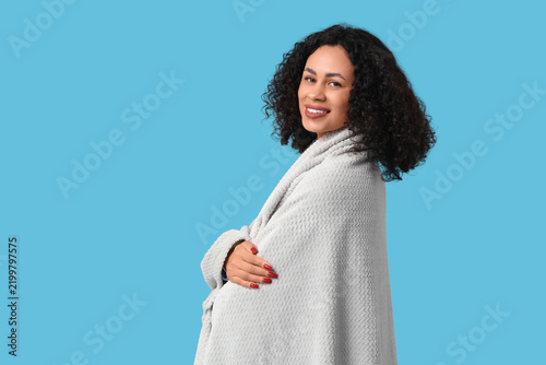 Beautiful young African-American woman with cozy blanket on blue background