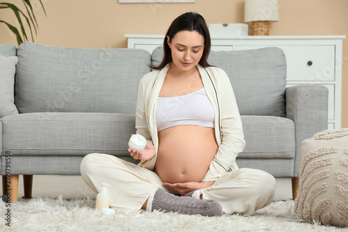 Beautiful young pregnant woman with jar of cosmetic cream sitting on carpet i...